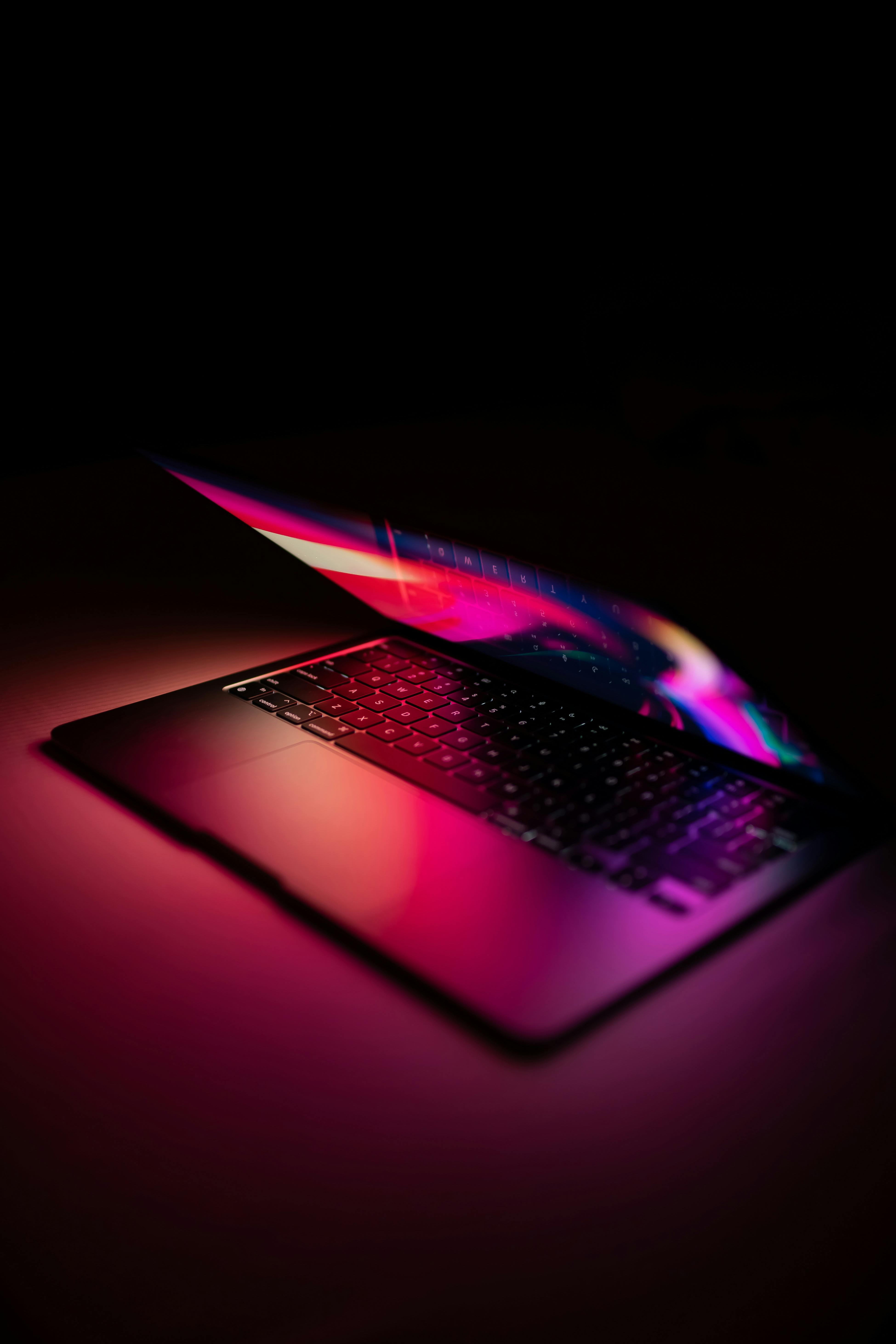 A sleek laptop partially open in a dark environment, with vibrant magenta and blue light reflecting off its surface and keyboard